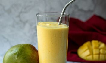 mango lassi smoothie in a glass with a straw and fresh mango next to it