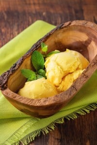 Homemade mango ice cream with fresh mint in olive wood bowl, served on green textile napkin over wooden textured background.