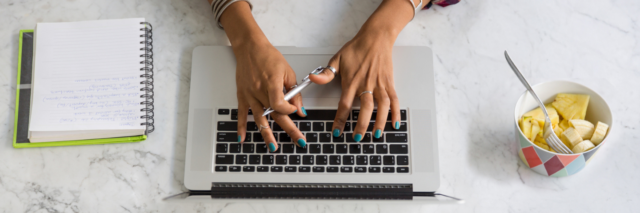 person's hands typing on computer with a bowl of pineapple and notebook with writing in it