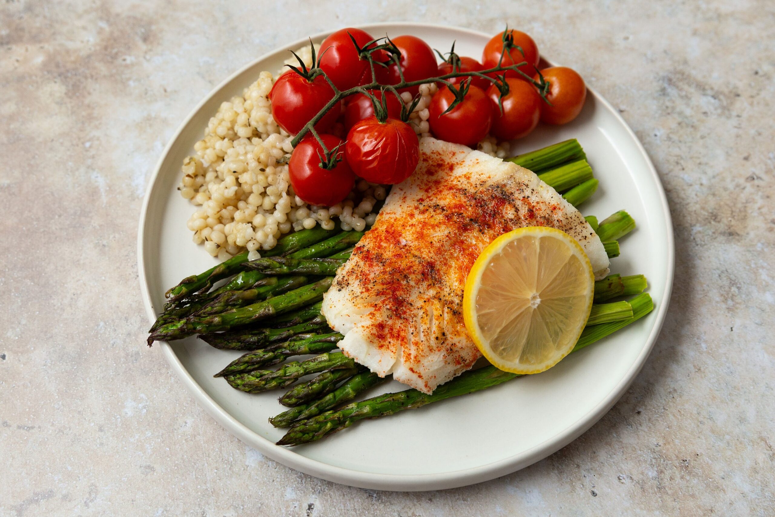 flaky spiced cod on top of roasted asparagus with couscous and cherry tomatoes on the side